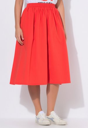 Frau trägt einen knielangen, leuchtend roten Rock mit elastischem Bund und weiße Turnschuhe, steht vor weißem Hintergrund.