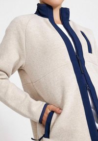 Beige Fleecejacke mit hohem marineblauen Kragen, frontaler Reißverschluss, kontrastierendem marineblauen Besatz, seitlichen Taschen und lässiger Passform.