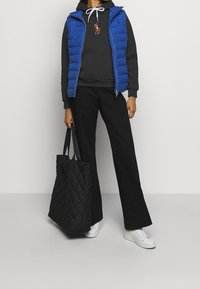 Chaleco inflable azul sobre una sudadera gris, pantalones negros de campana y zapatillas blancas. Bolso tote de cuero acolchado negro. Detalle de cremallera en el chaleco.