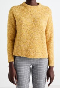 Texturerad gul stickad tröja med rund halsringning och långa ärmar, ihop med grå och svart rutiga byxor. Mjuk, fluffig yta.