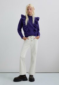 Suéter de punto morado con acentos con volantes, combinado con jeans blancos de corte acampanado y botas negras robustas. Textura suave, diseño moderno y colores distintivos.