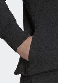 Main d'une personne partiellement dans la poche d'un vêtement à manches longues gris foncé avec des détails de couture visibles.