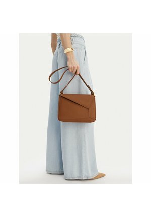 Mujer con vaqueros de pierna ancha de color azul claro, sosteniendo un bolso de cuero marrón por la correa, sobre un fondo liso.