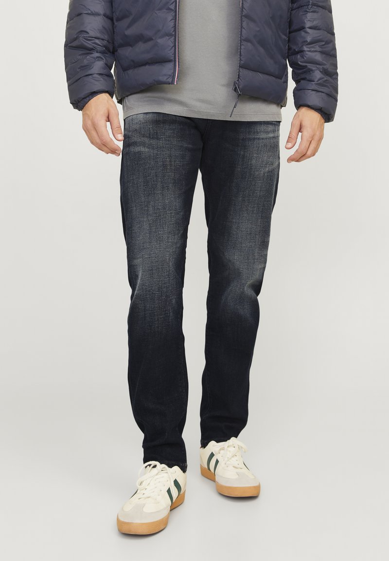 Temno modri jeans s ožjim krojem, z blagim vintage učinkom, dokončane hlačnice in vidnim šivanjem. Ujemajo se z belimi supergami.