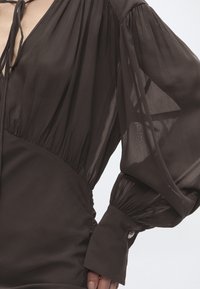 Διάφανο μακρύ μανίκι με φαρδύ μανσέτα και λεπτομέρεια κουμπιού σε σκούρο καφέ φόρεμα με σφιχτή μέση και κλείσιμο με κορδόνι σε σχήμα V.