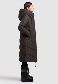 Langer, dunkelbrauner Daunenmantel mit hohem Kragen und Chevron-Muster. Hat Seitentaschen und einen mit Reißverschluss verstellbaren Saum. Schwarze Schnürstiefel.