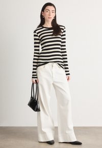 Top de rayas blanco y negro de manga larga combinado con pantalones anchos blancos. La modelo sostiene un pequeño bolso negro y lleva zapatos de punta.