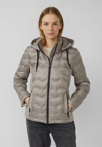 Vrouw in een lichtgrijze gewatteerde jas met capuchon, zwarte rits en zakken, een beige trui en een donkere spijkerbroek, staand tegen een effen achtergrond.
