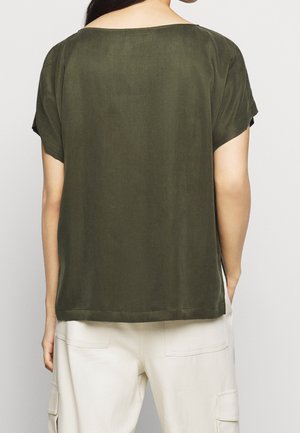 Kobieta ubrana w oliwkowo-zieloną, luźną bluzkę z krótkim rękawem oraz kremowe spodnie cargo, pokazana od tyłu na jednolitym tle.