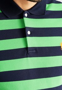 Pikétröja med gröna och marinblå horisontella ränder, med krage, tre knappar och en liten orange hästlogo på bröstet.