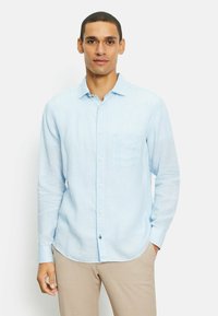 Camicia azzurra a maniche lunghe in tessuto di lino, con taschino sul petto e orlo arrotondato, abbinata a pantaloni beige.