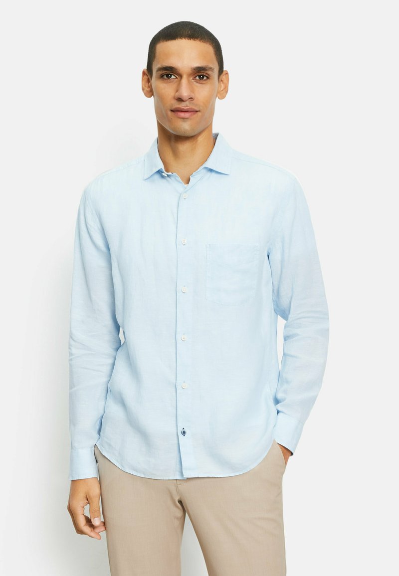 Lichtblauwe blouse met lange mouwen in linnen stof, met een borstzak en een ronde zoom, gecombineerd met beige broeken.