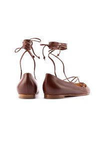 Le ballerine in pelle marrone presentano lacci incrociati alla caviglia, una punta arrotondata e una suola di legno piatta. La texture è liscia con dettaglio a intaglio.