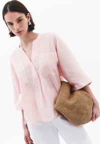 Blusa a righe rosa chiaro con scollo a V, due tasche frontali e maniche a tre quarti, abbinata a una borsa di paglia intrecciata.
