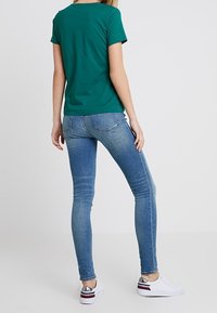 Mujer con una camiseta verde de manga corta, jeans ajustados azul claro y zapatillas blancas con acentos en plata y rojo, de pie mirando hacia atrás.