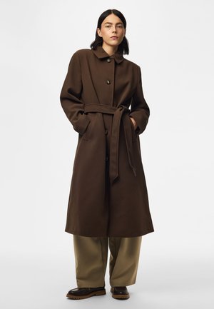 Femme debout portant un long manteau marron foncé ceinturé avec des boutons, un pantalon beige à jambes larges et des chaussures marron foncé sur un fond blanc uni.