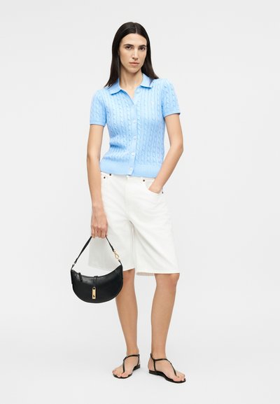 Sieviete, kas valkā gaismazilu īsroku adītu topu un baltas ceļos garas šortus, tur melnu rokassomu un valkā melnas sandaletes.