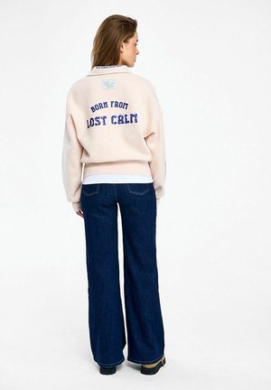 Kobieta stojąca tyłem, ubrana w jasnoróżową bluzę z napisem "BORN FROM LOST CALM" oraz ciemnoniebieskie, szerokie dżinsy.