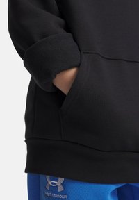 Main dans la poche de sweat-shirt noir avec poignet en polaire retroussé, au-dessus d'un pantalon bleu Under Armour avec logo visible.