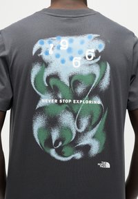 Πίσω μέρος σκουρόχρωμου γκρι μπλουζακιού με μπλε και πράσινο αφηρημένο σχέδιο, λευκούς αριθμούς, το κείμενο "NEVER STOP EXPLORING" και το λογότυπο The North Face.