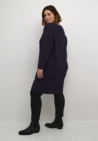 Purple ribbed long-sleeve dress with a relaxed fit, features a round neckline and side slits. Paired with black fitted pants and ankle boots.