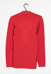 Červené sportovní tričko s dlouhým rukávem a hladkou texturou, s designem síťové panely na zadní straně a kulatým výstřihem. Visí na černém háčku.
