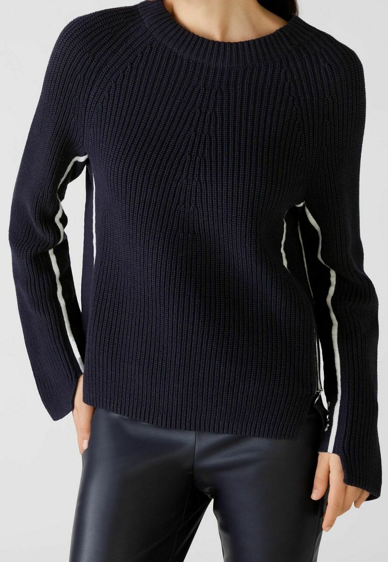 Pull en maille bleu marine avec texture côtelée, col rond et bandes blanches sur les côtés ; doté de manches longues et de fermetures éclair latérales.