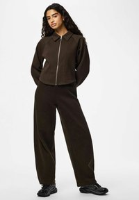 Veste marron à fermeture éclair avec col et manches longues, associée à un pantalon marron à jambes larges. Les deux sont en tissu doux, portés avec des baskets noires.