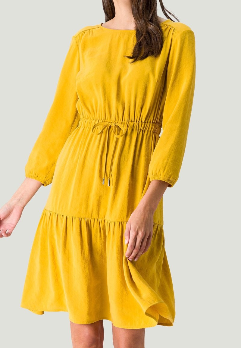 Femme portant une robe jaune moutarde à manches longues, longueur genou, avec taille froncée et lien à coulisser.