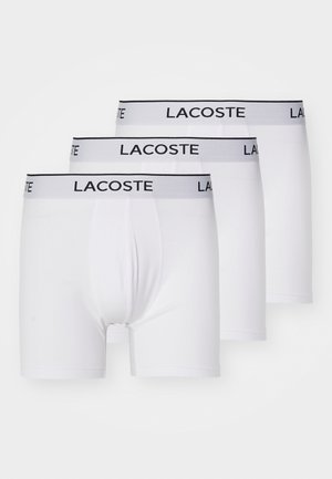 Boxers de algodón blancos en un paquete de tres, con una cinturilla gris que presenta la marca "LACOSTE" en negro. Textura suave, corte clásico.