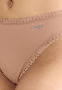 Beige bikini i bomuldsblanding med blonderkant ved taljen, med en lyseblå "sloggi"-logo på fronten. Glat tekstur og minimal dækning.