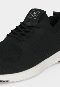 Baskets en maille noire avec lacets noirs et semelle blanche, avec une étiquette noire avec texte blanc "M O P" sur la languette.