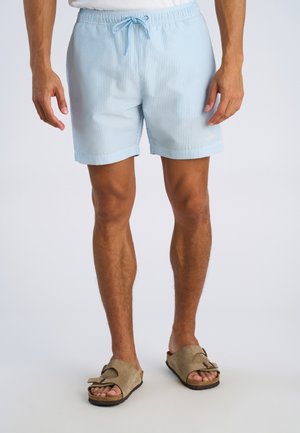 Costume da bagno a righe azzurre e bianche con vita elastica, coulisse e tasche laterali, abbinato a sandali beige a ciabatta.