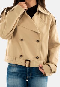 Donna che indossa una giacca corta doppiopetto beige con grandi bottoni e cintura, sopra jeans blu, mentre aggiusta il colletto con una mano.