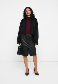 Sieviete stāv, ģērbjusies melnā īsajā jakā, bordo turtleneckā, melnā ādas vidusgaruma svārkos ar priekšējo spraugu, melnās augstpapēžu kurpēs un turēdama melnu roku somu.