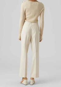 Femme portant un pantalon large à taille haute de couleur crème, un pull à manches longues en ribbed beige, et des sandales compensées beiges, se tenant de dos.