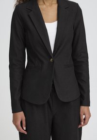 Blazer noir en tissu texturé, avec un seul bouton, des revers crantés et deux poches avant, porté sur un haut blanc.