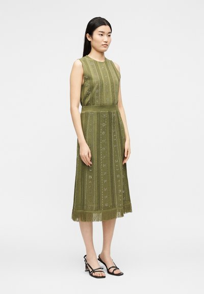 Γυναίκα στέκεται φορώντας αμάνικο μεσομπούστο ελιάς με κάθετα σχέδια και κρόσσια στο τελείωμα, συνδυασμένο με μαύρα σανδάλια με τακούνι.