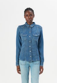 Camicia in denim con colletto classico, due tasche sul petto e chiusura con bottoni. È di un blu medio con una texture liscia.