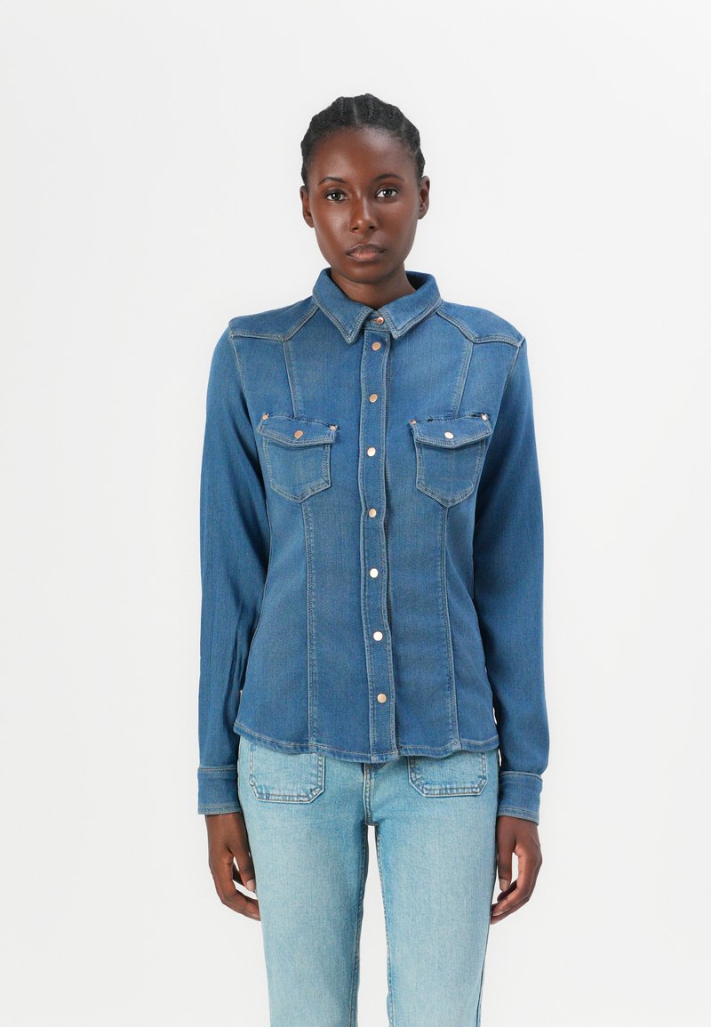 Camicia in denim con colletto classico, due tasche sul petto e chiusura con bottoni. È di un blu medio con una texture liscia.