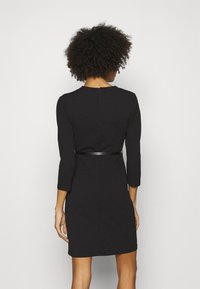 Žena s kudrnatými vlasy, oblečená v přiléhavých černých šatech s rukávy po tři čtvrtiny a tenkým černým páskem, stojí čelem k jednoduché světlé zdi.