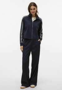 Marineblauw trainingspak met een ritsjacks met witte strepen en wijde pijpen. Gladde textuur, casual pasvorm en zijzak op het jack.