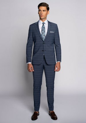 Dopasowany niebieski garnitur w kratę, jednorzędowa marynarka, klapy w stylu notch, dopasowane spodnie, biała koszula elegancka, wzorzysty krawat, brązowe skórzane buty.