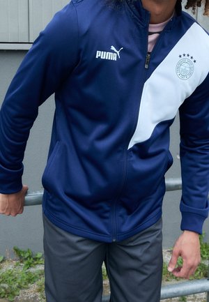 Person trägt eine marineblaue und weiße Puma-Zipjacke mit einem Sportclub-Logo auf der Brust und lehnt sich draußen an ein Metallgeländer.