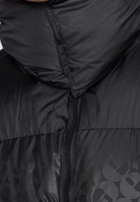 Doudoune noire avec un col montant, fermeture éclair, matériau brillant et une texture à motifs subtils sur le tissu.