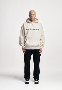 Beige hooded sweatshirt with "2Y STUDIOS" printed in black, paired with black pants and black-and-white sneakers. Plain background.