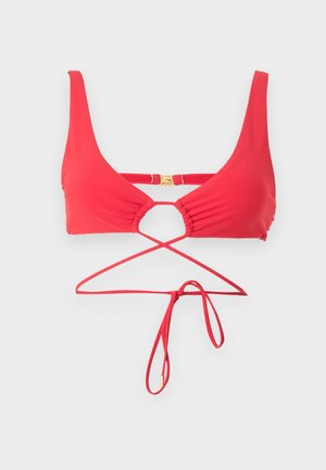 Kobieta z mokrymi włosami, ubrana w czerwone bikini z przewlekanymi ramiączkami, stojąca na jednolitym białym tle, patrząca w kamerę.