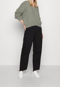 Grüner Wollpullover mit V-Ausschnitt, schwarze, hoch taillierte Hose mit Knopfleiste und weiße Turnschuhe vor neutralem Hintergrund.