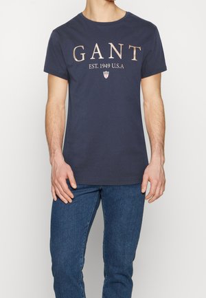 Mężczyzna w granatowej koszulce GANT z napisem "EST. 1949 U.S.A" i emblematem, w połączeniu z niebieskimi dżinsami, stojący na tle białym.