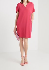Kurzes, knieumspielendes, pinkes Kleid mit V-Ausschnitt, strukturiertem Stoff und seitlichen Taschen, kombiniert mit einer pinken Clutch und passenden Sandalen.